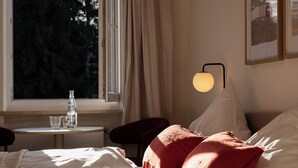 Een netjes opgemaakt bed met rode kussens, een nachtkastje met een fles en glazen, en een raam met gordijnen.