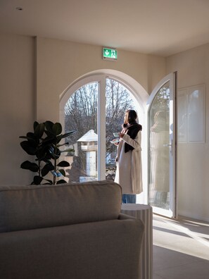 A woman standing by a large arched window in a room with a plant, a coat rack, and a door with a glass panel. - Lindenhof (Siegsdorf)