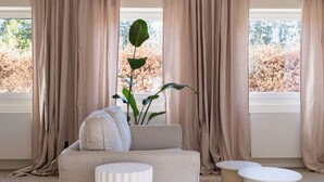 Een moderne woonkamer met een beige bank, twee witte bijzettafeltjes en een groot raam met beige gordijnen.