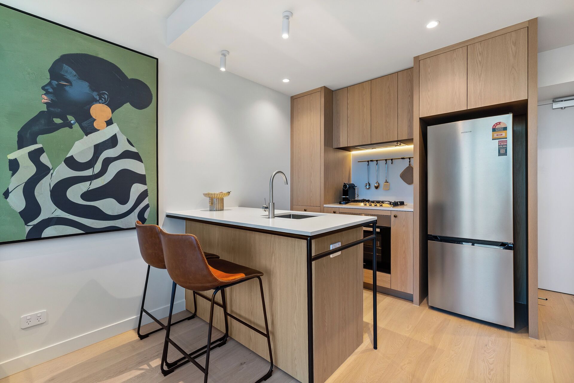 A modern kitchen with wooden cabinets, a stainless steel refrigerator, a bar stool, and a painting on the wall.