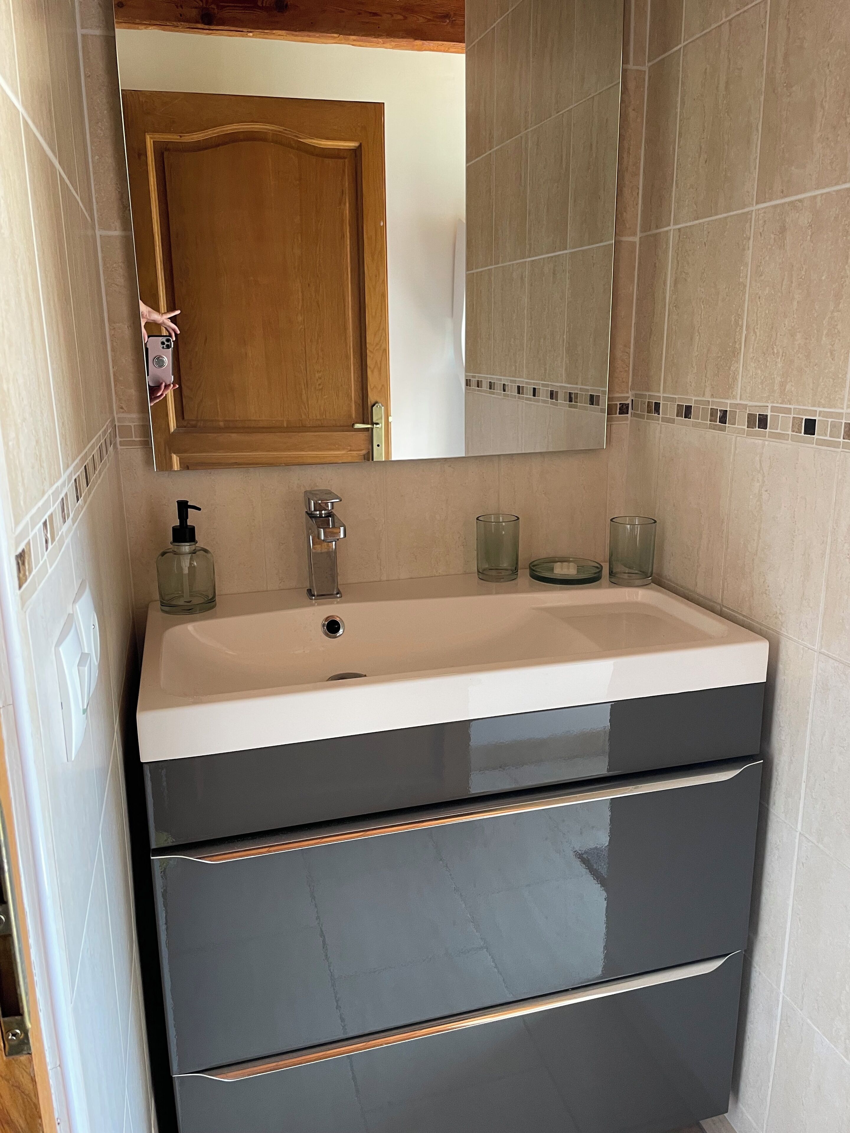 Une salle de bain avec une porte en bois, un lavabo blanc avec un robinet noir et un miroir au-dessus.