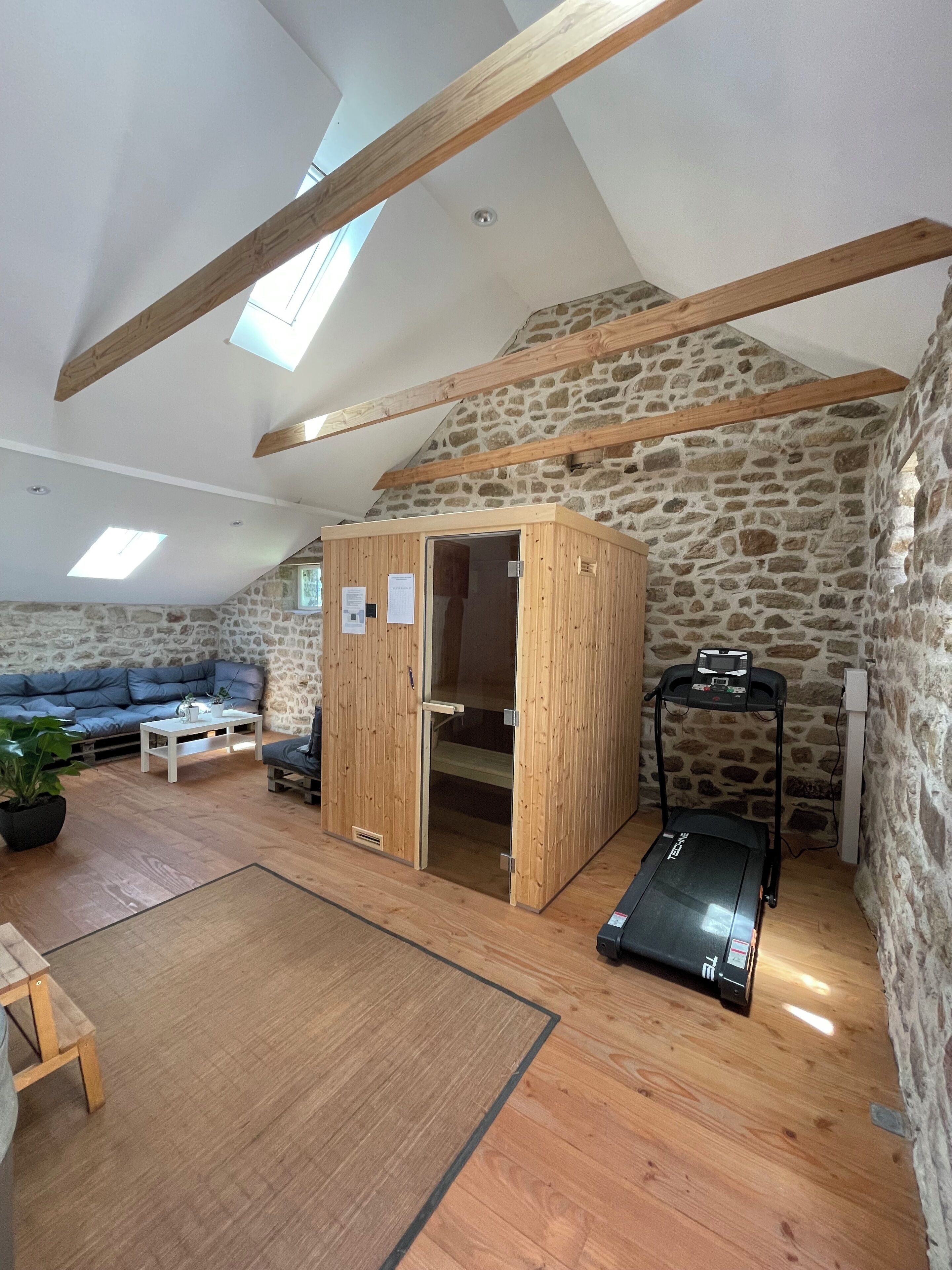 Una habitación con pared de piedra, vigas de madera, una cinta de correr, un armario de madera, un sofá y una pequeña mesa.