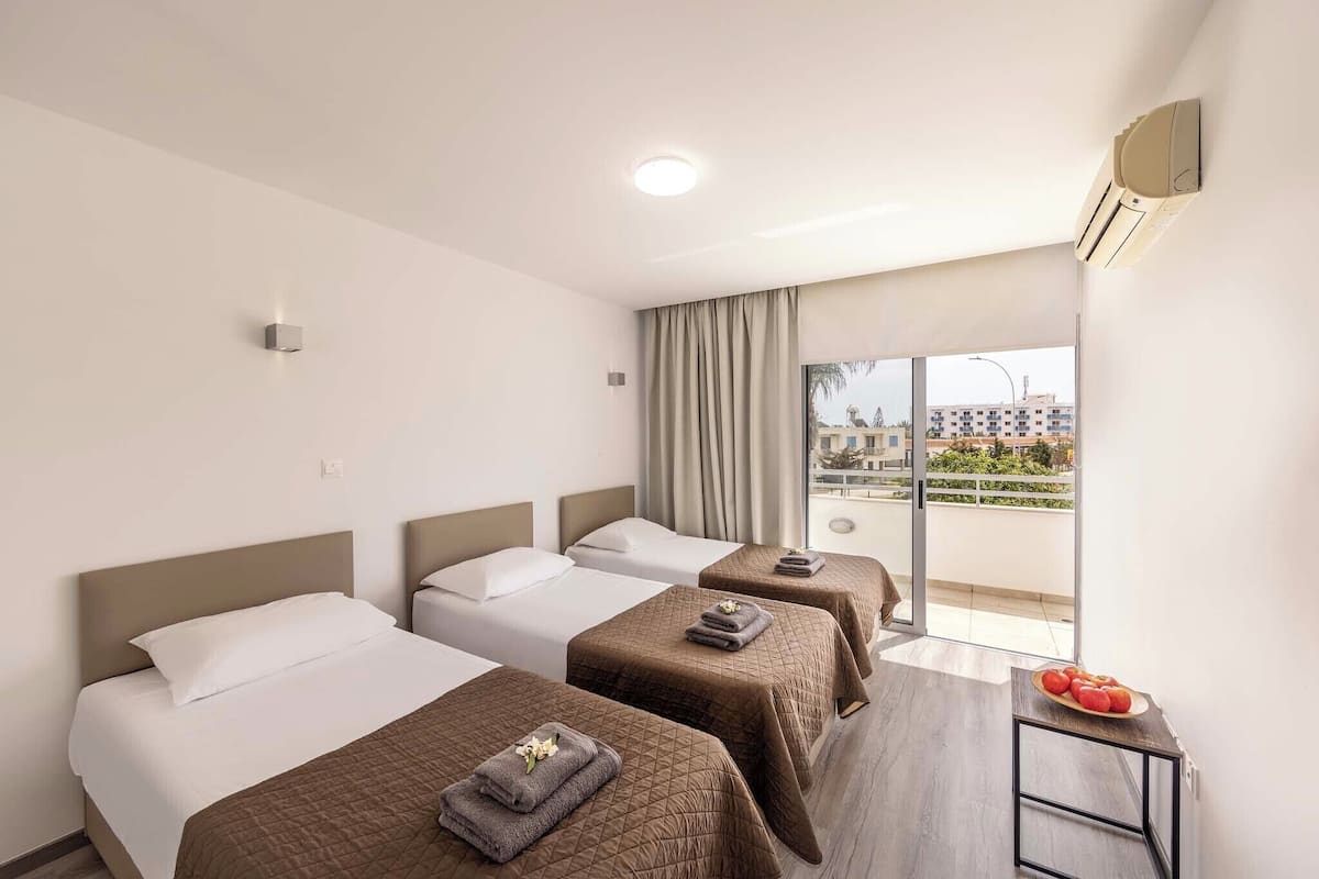 A hotel room with three beds, a small table with a fruit bowl, and a view of the city through large windows.