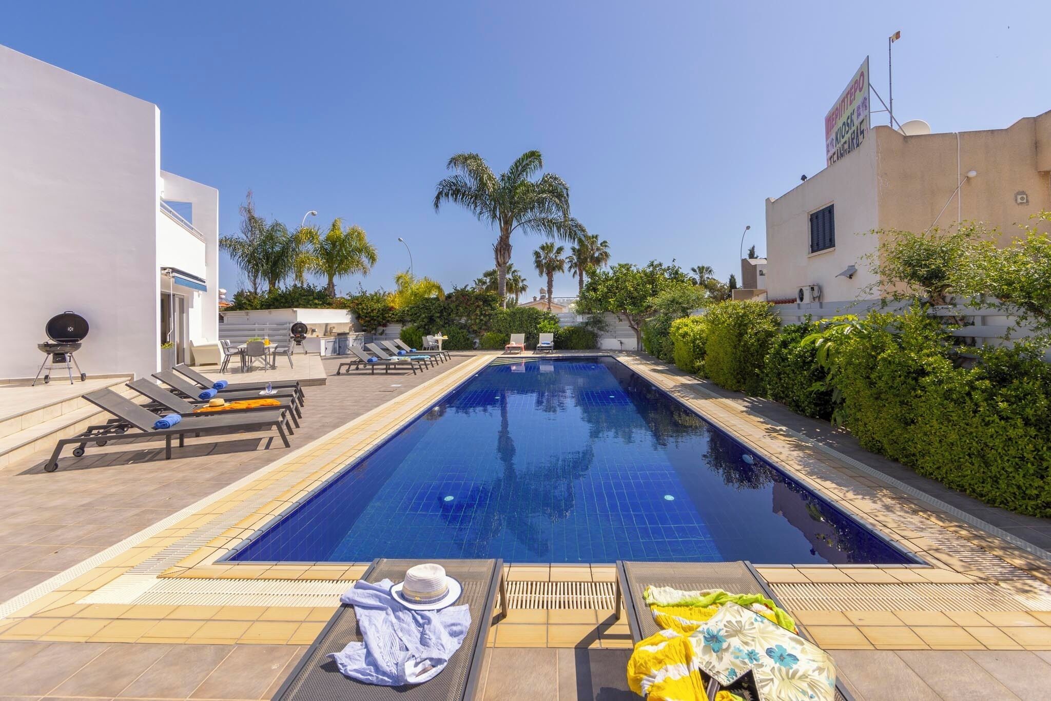 Piscina con sillones de descanso y sombrillas, rodeada de vegetación y edificios.