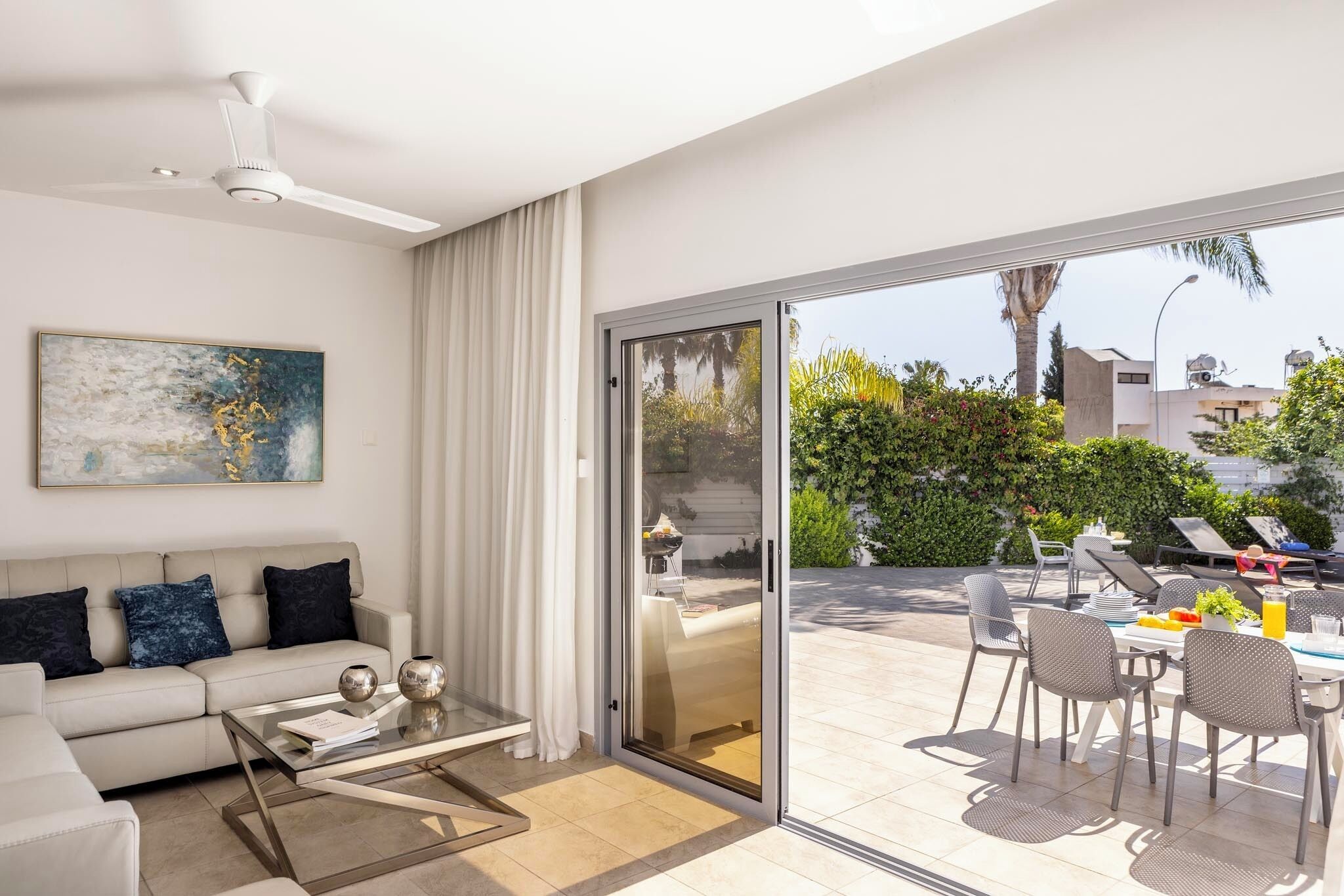 Uma sala de estar moderna com sofá, mesa de centro e uma porta de vidro deslizante que dá para uma área de jantar ao ar livre com cadeiras e mesa.