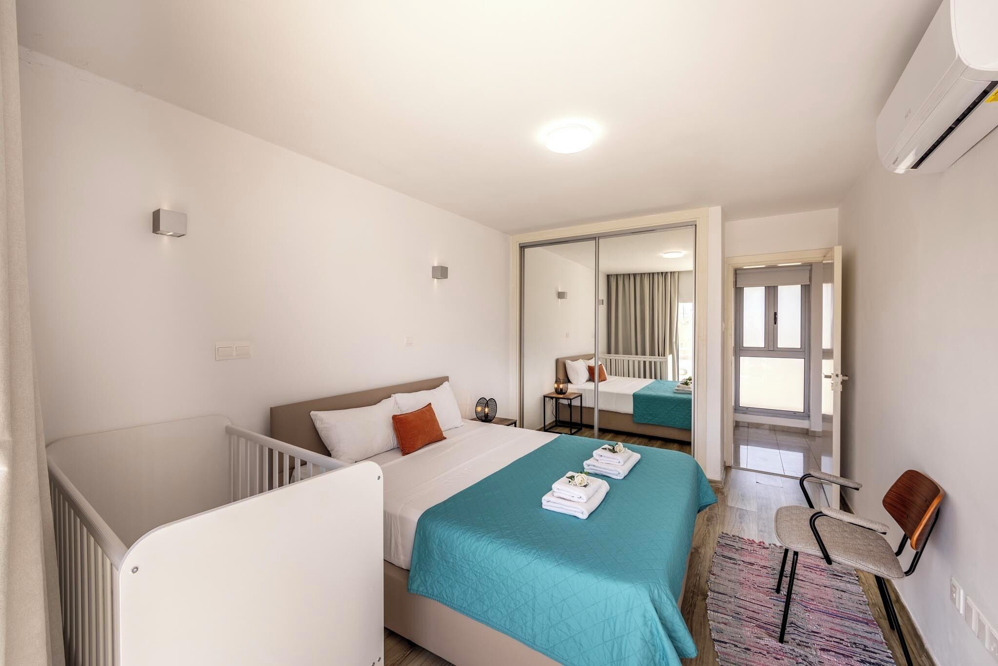 A modern hotel room with a large bed, a separate crib, a mirror, a small table, and a chair.