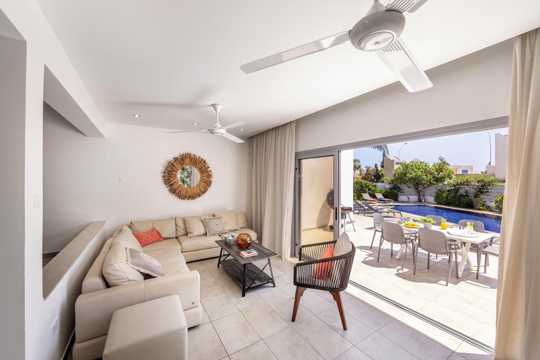Un soggiorno moderno con un divano, un tavolino da caffè e un'area pranzo con sedie e tavolo. Si nota una ventola a soffitto, un elemento decorativo a parete e vista su una piscina esterna.