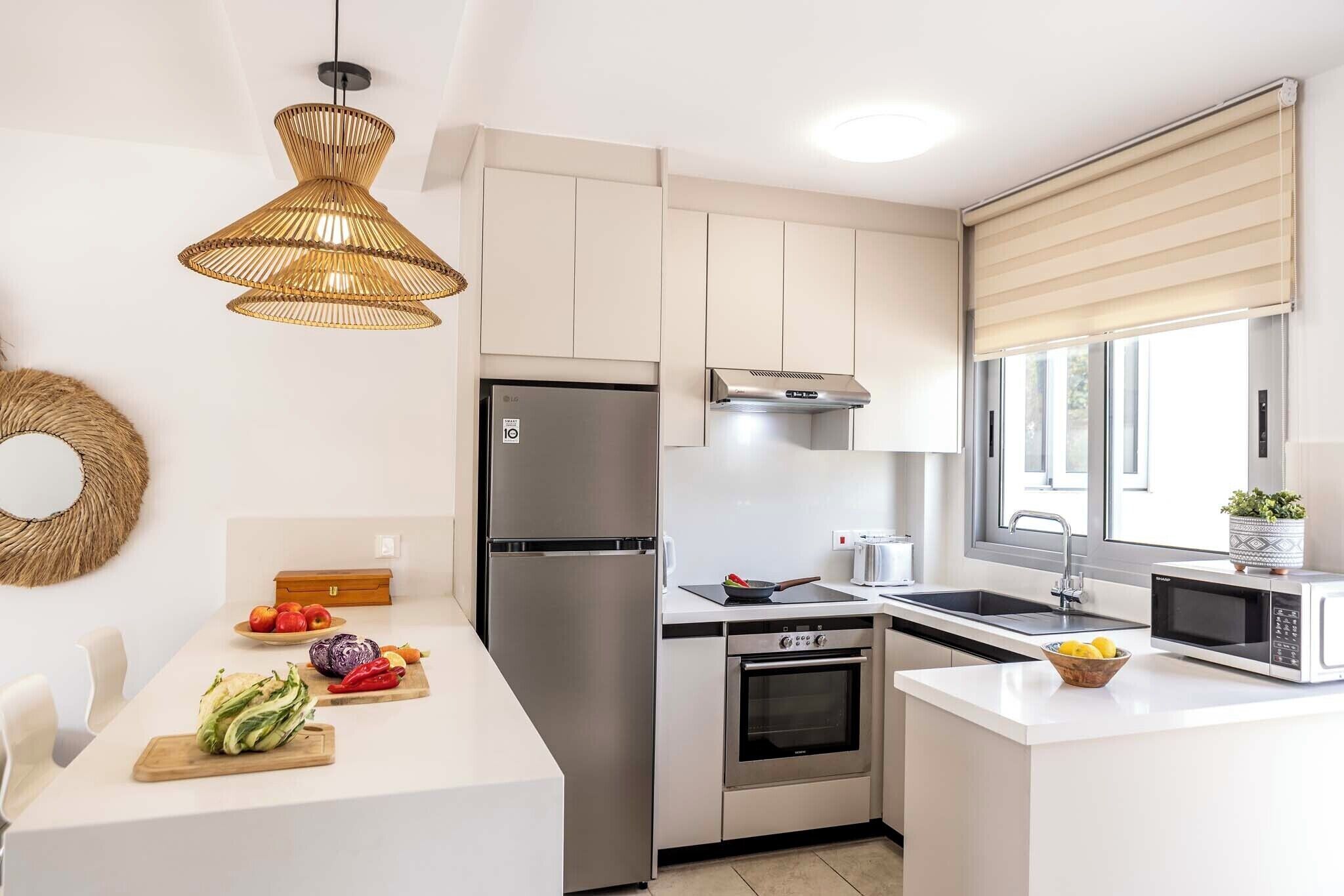 A modern kitchen with white cabinetry, stainless steel appliances, a dining area with a white table, and a large window with blinds.