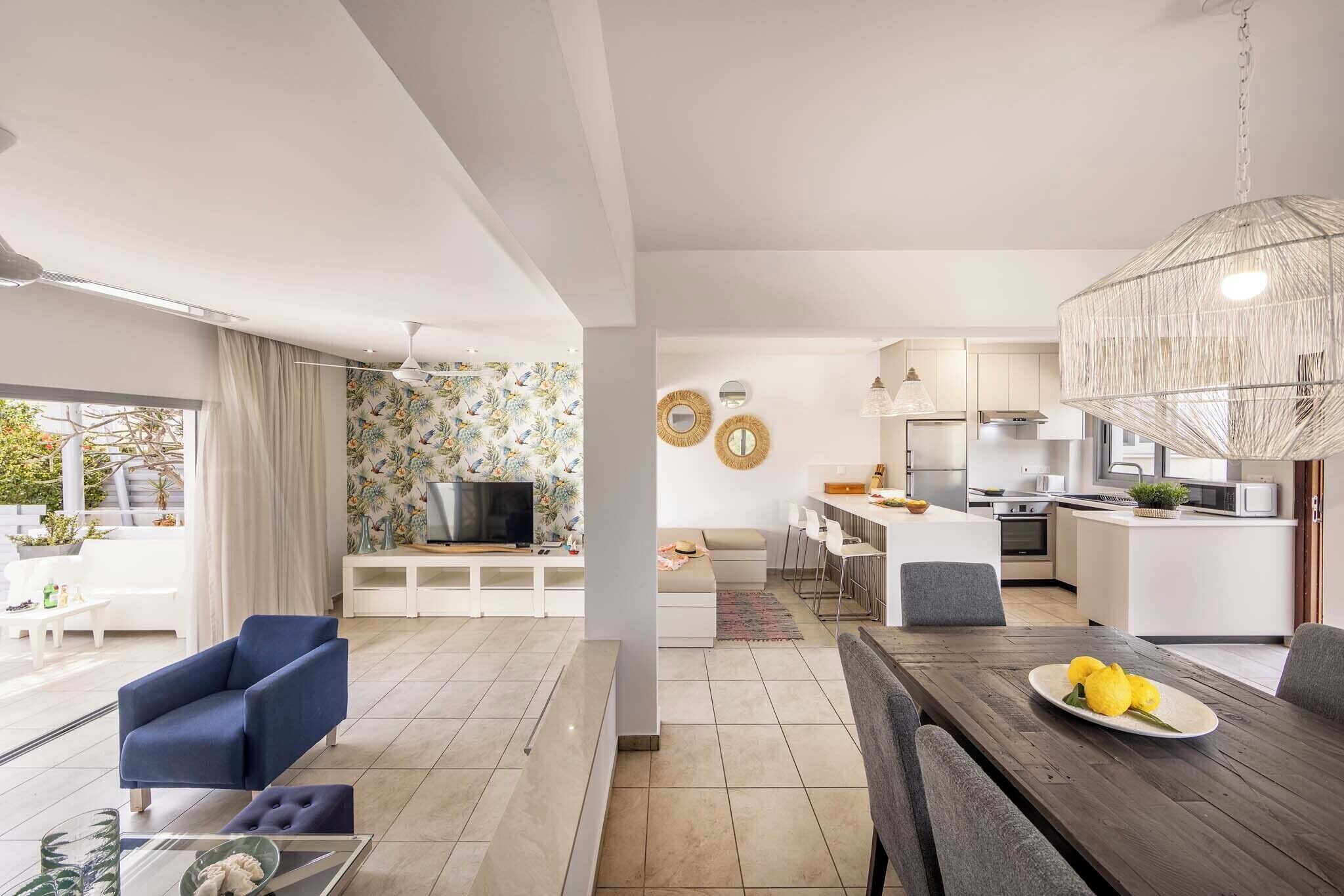 A modern living room with a sofa, a coffee table, and a dining table with a plate of lemons. There is a kitchen area with a dining set and a TV in the background.