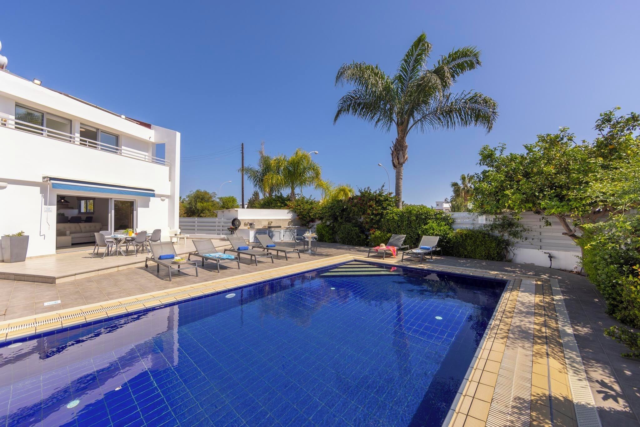 Un área de piscina moderna con una piscina azul cristalina, sillones de descanso y un edificio blanco con balcón.