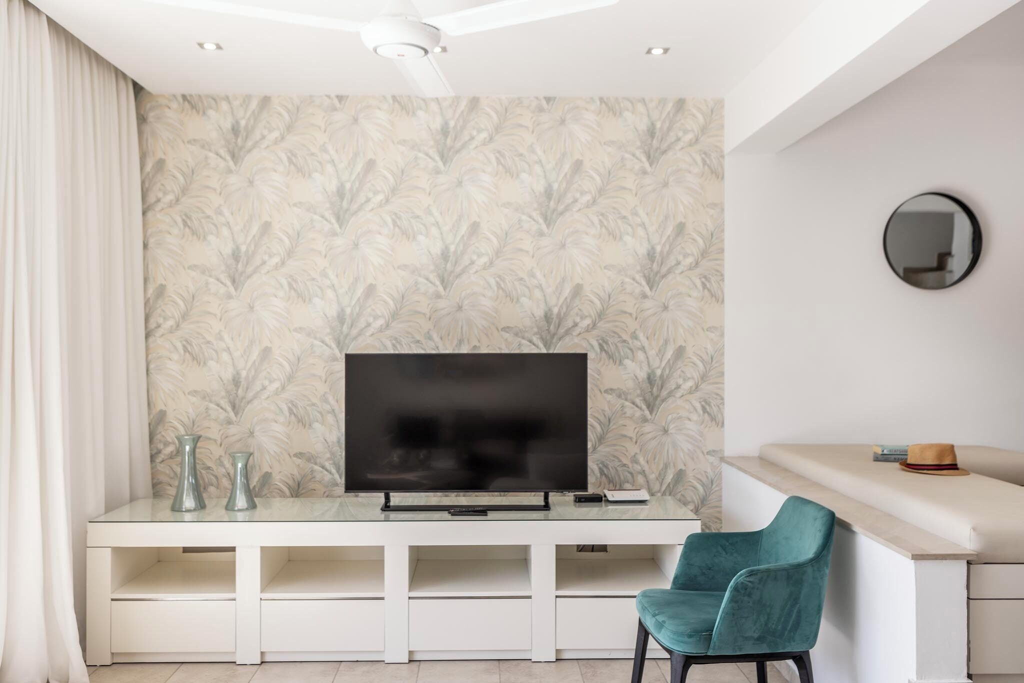 Un soggiorno moderno con una TV a schermo piatto su un mobile bianco, una poltrona color petrolio e carta da parati decorata.