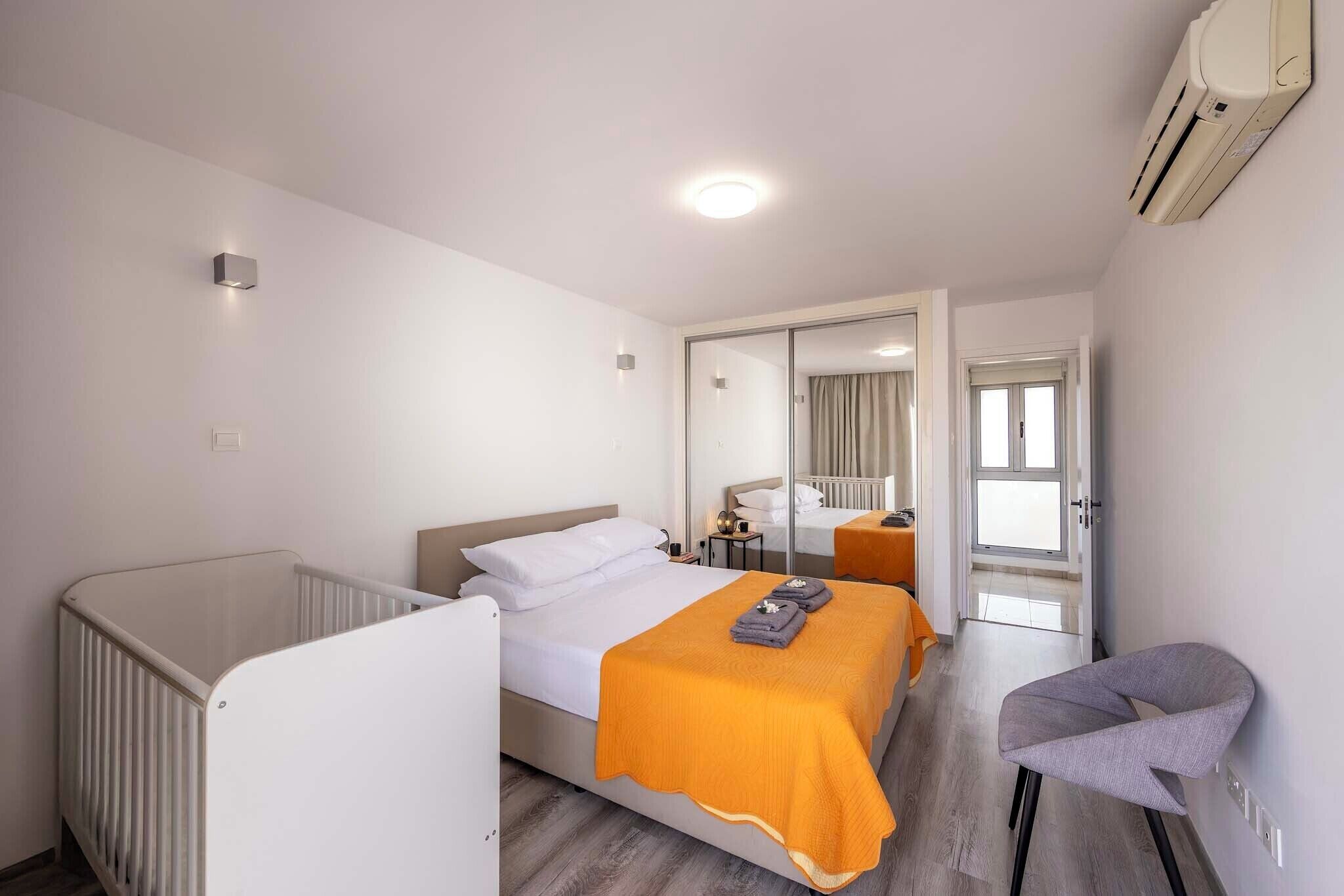 A modern hotel room with a single bed, a crib, a chair, and a large mirror.