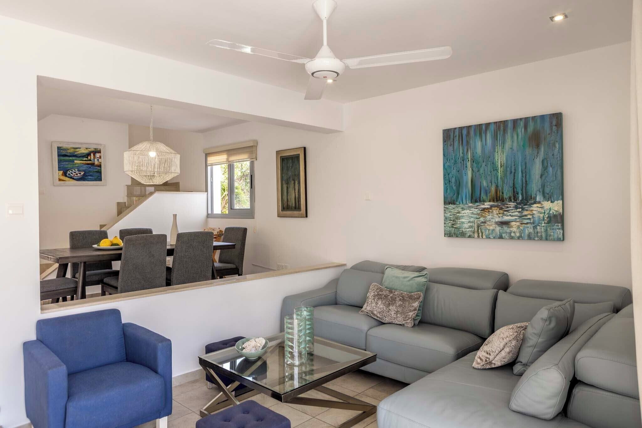 A modern living room with a grey sofa, blue armchair, and a glass coffee table. A ceiling fan and artwork are visible on the walls.