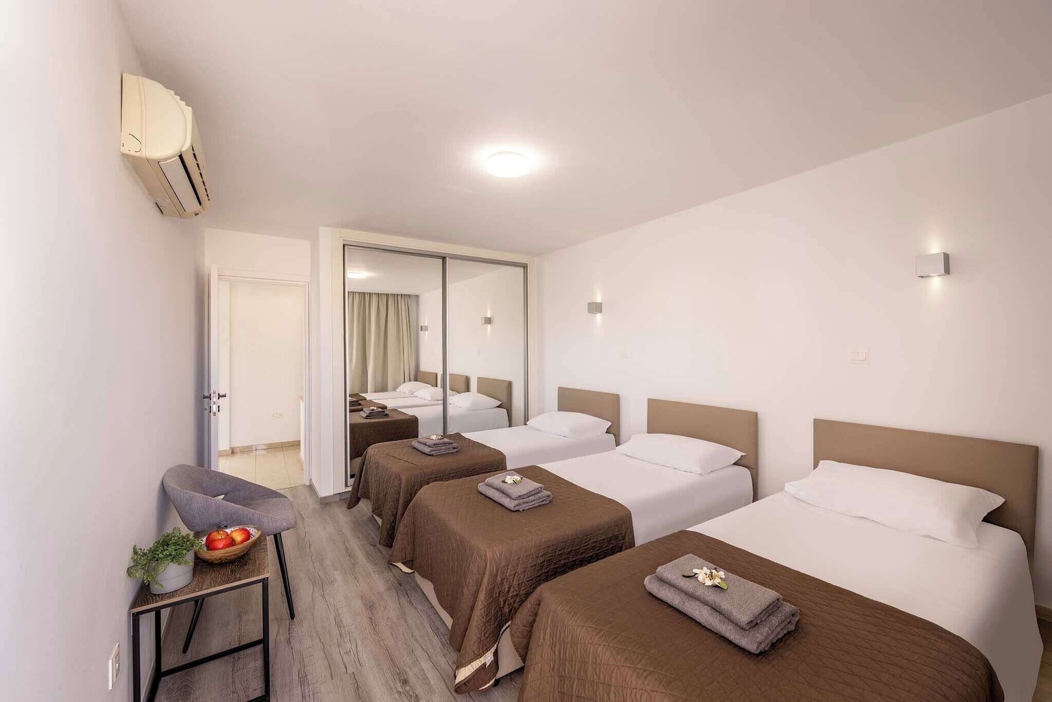 A hotel room with three single beds, a small table with a plant and fruit, and a chair.