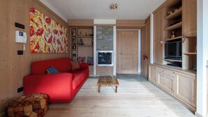 Een woonkamer met een rode bank, een klein houten salontafeltje en een stenen openhaard. Er staat een televisie op een houten kast en een kleurrijk abstract schilderij aan de muur.