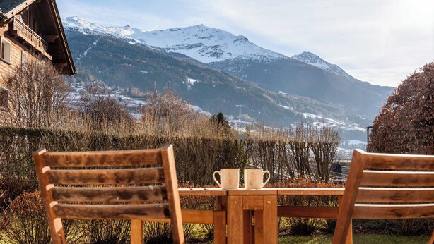 Een houten tafel met twee stoelen en een klein tafeltje, met uitzicht op een besneeuwd berglandschap met een kerk in de verte.