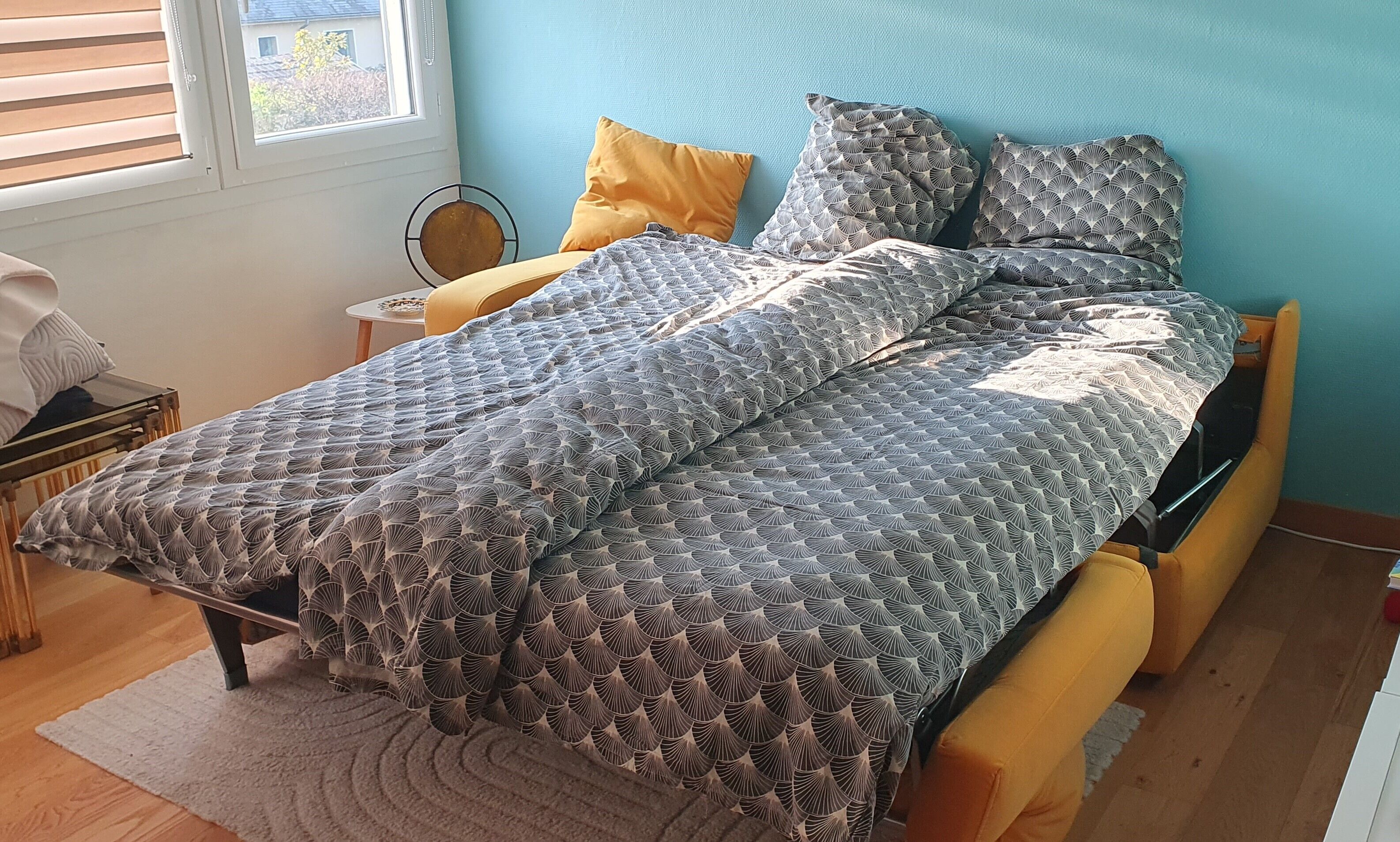 A neatly made bed with a patterned duvet, a small bedside table with a lamp, and a window with blinds.