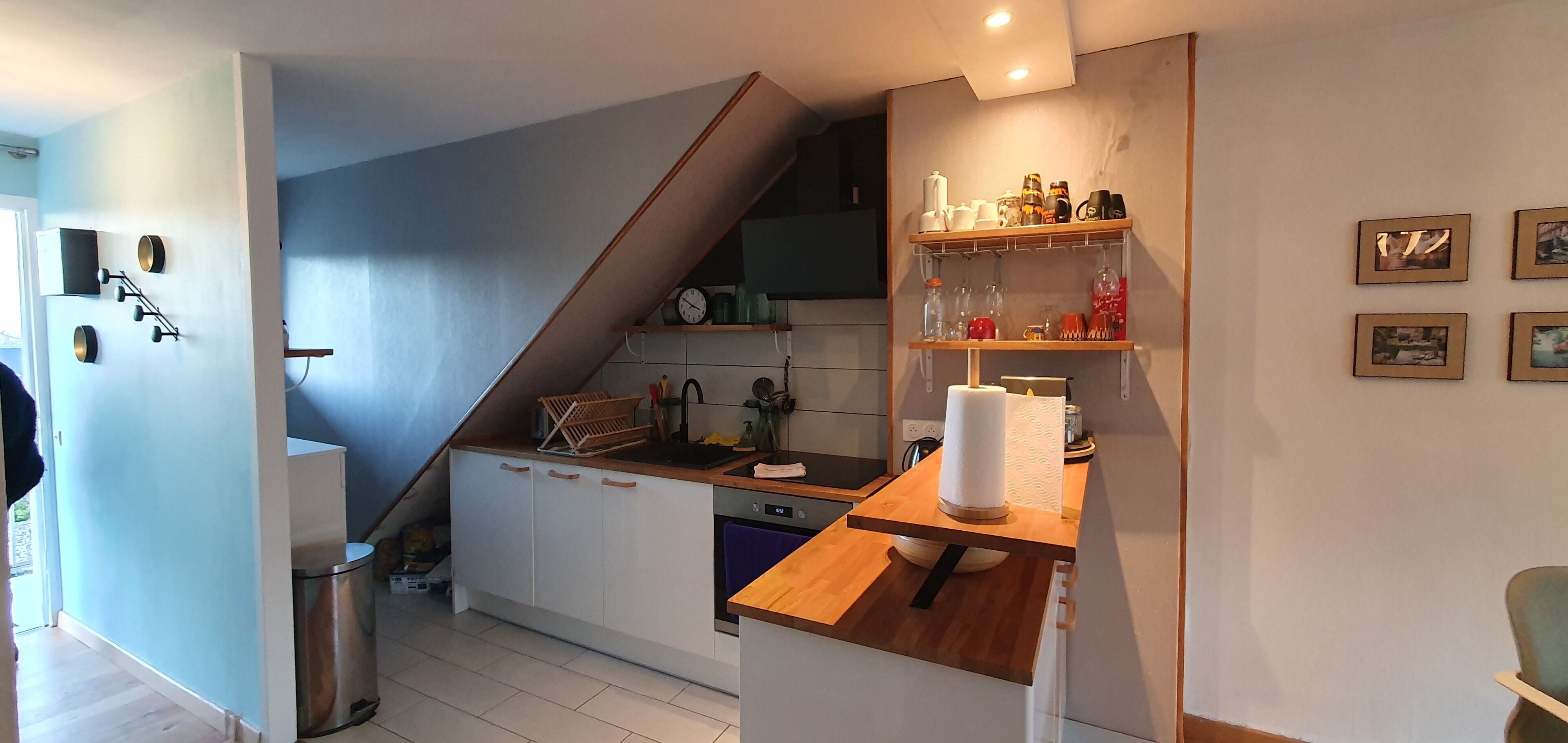 Eine kompakte Küche mit weißen Schränken, einer Holz Arbeitsplatte und einem eingebauten Backofen. Eine Treppe führt auf eine höhere Ebene, und an der Wand befindet sich ein Regal mit Küchenutensilien und gerahmten Bildern.