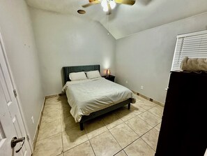 Un dormitorio con cama, mesita de noche, ventilador de techo y ventana con persianas.