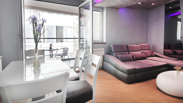 Un salon moderne avec une table à manger en verre, des chaises blanches, un canapé d’angle et une table basse ronde.