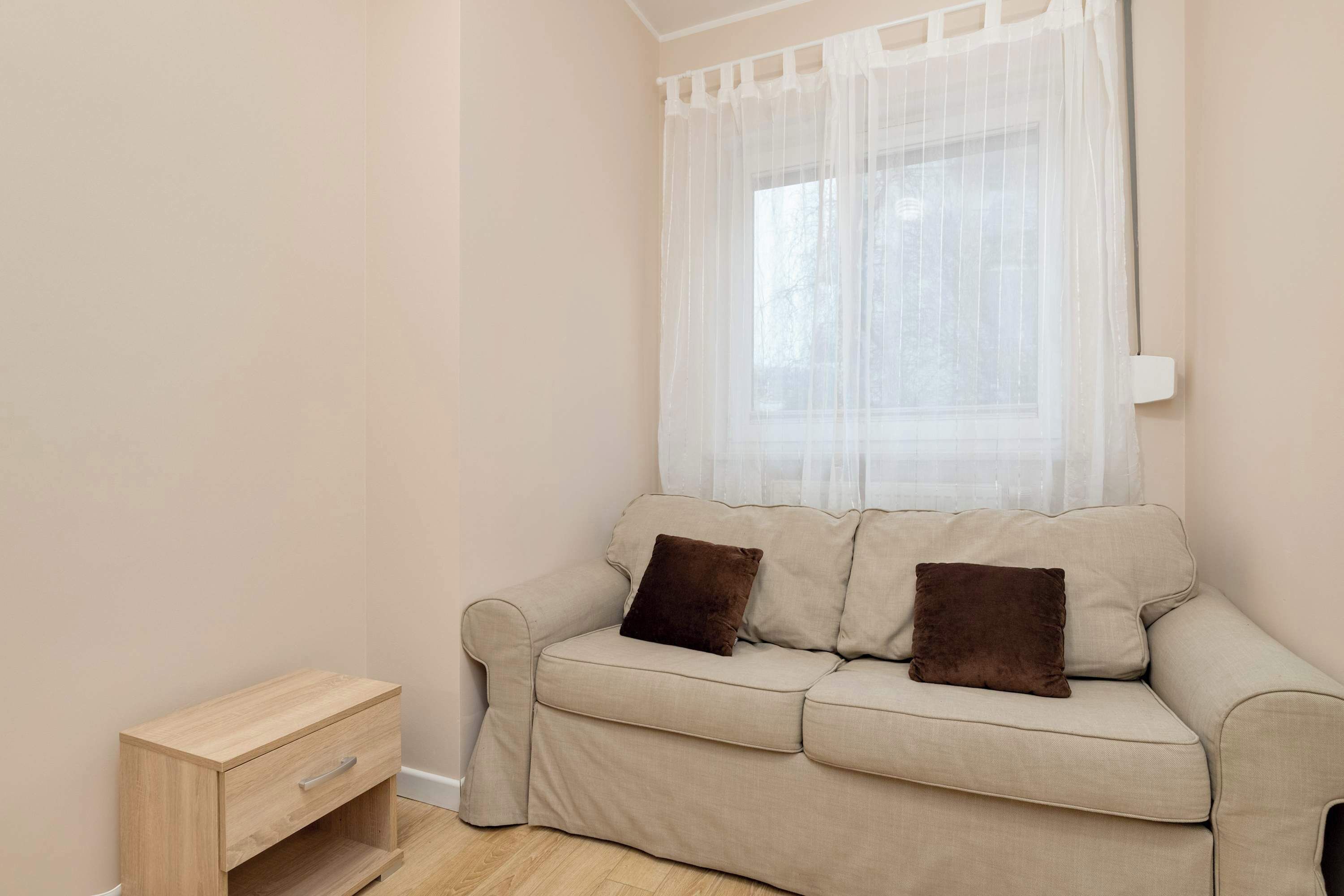 A beige sofa with two brown cushions, a small wooden side table, and sheer white curtains covering a window.