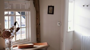 Et værelse med et træbord, en vase med tørrede blomster, et vindue med rullegardiner, et billede på væggen og en radiator.