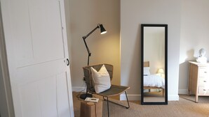 Et soveværelse med en hvid dør, en gulvlampe, en stol med pude, et stort spejl, en seng med hvid dyne og en kommode med en brystfigur.