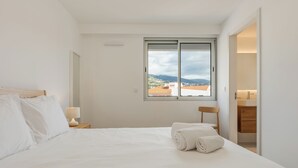 Un llit ben fet amb llençols blancs, una làmpada de nit i una vista d'un paisatge urbà a través d'una finestra.