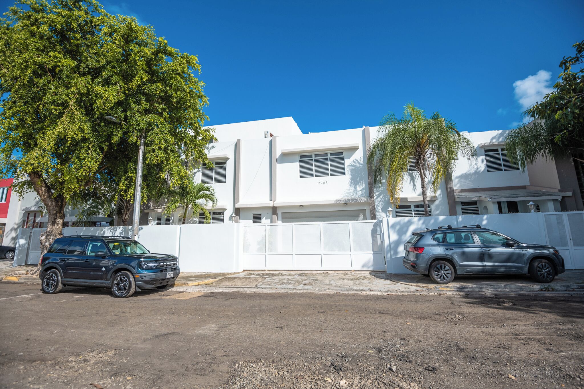 Un edificio de dos pisos con fachada blanca, un garaje y un árbol al frente.