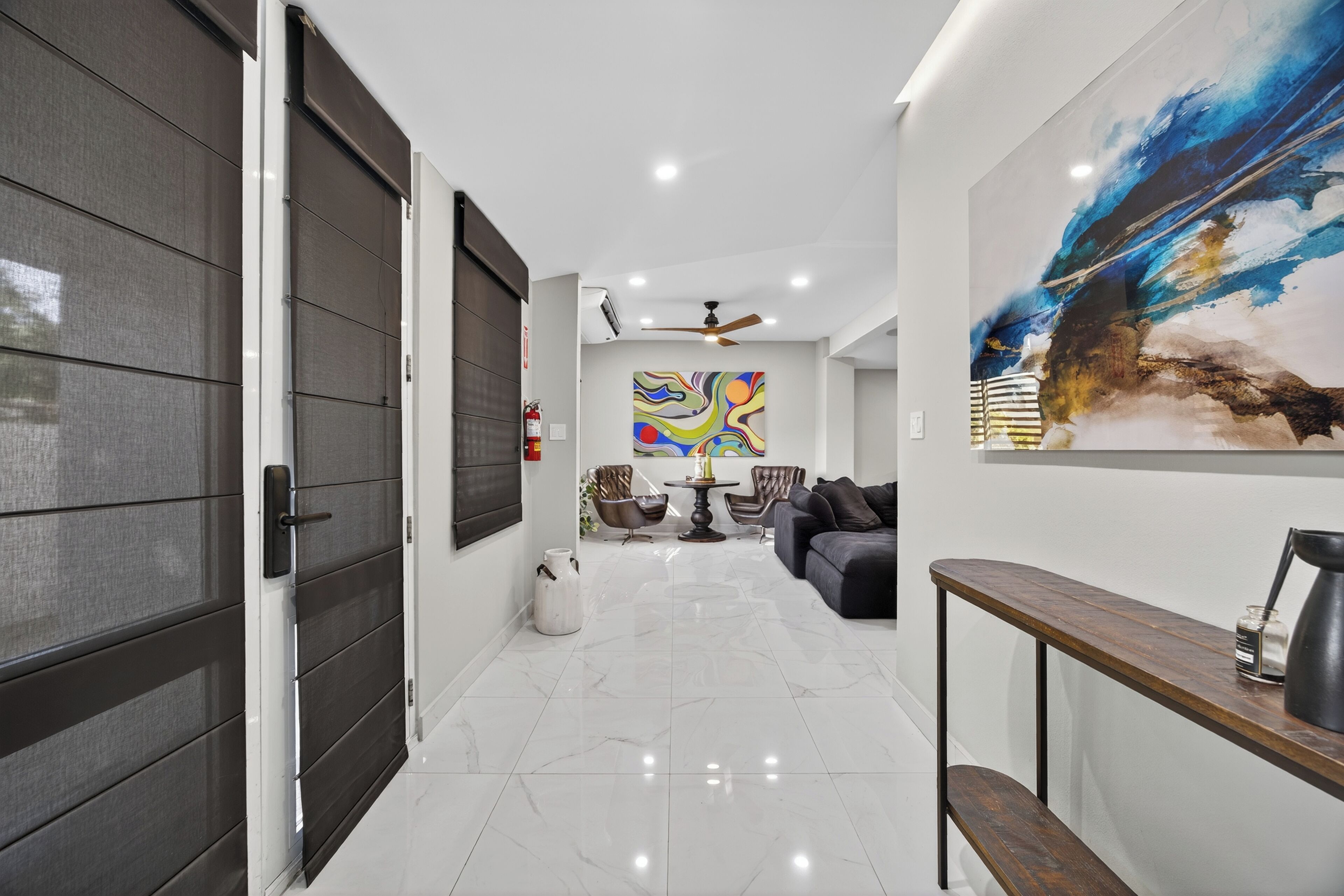 A modern interior with a hallway leading to a living area, featuring a large abstract painting, a ceiling fan, and a coffee table with chairs.