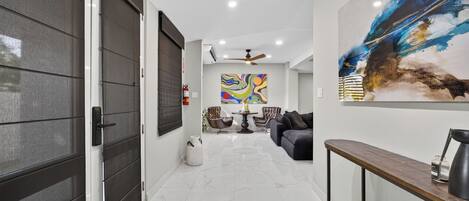 A modern interior with a hallway leading to a living area, featuring a large abstract painting, a ceiling fan, and a coffee table with chairs.