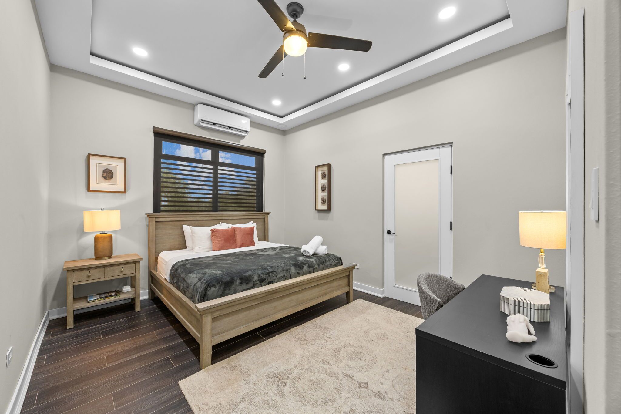 A bedroom with a bed, bedside tables, a desk, a chair, a ceiling fan, and a door.