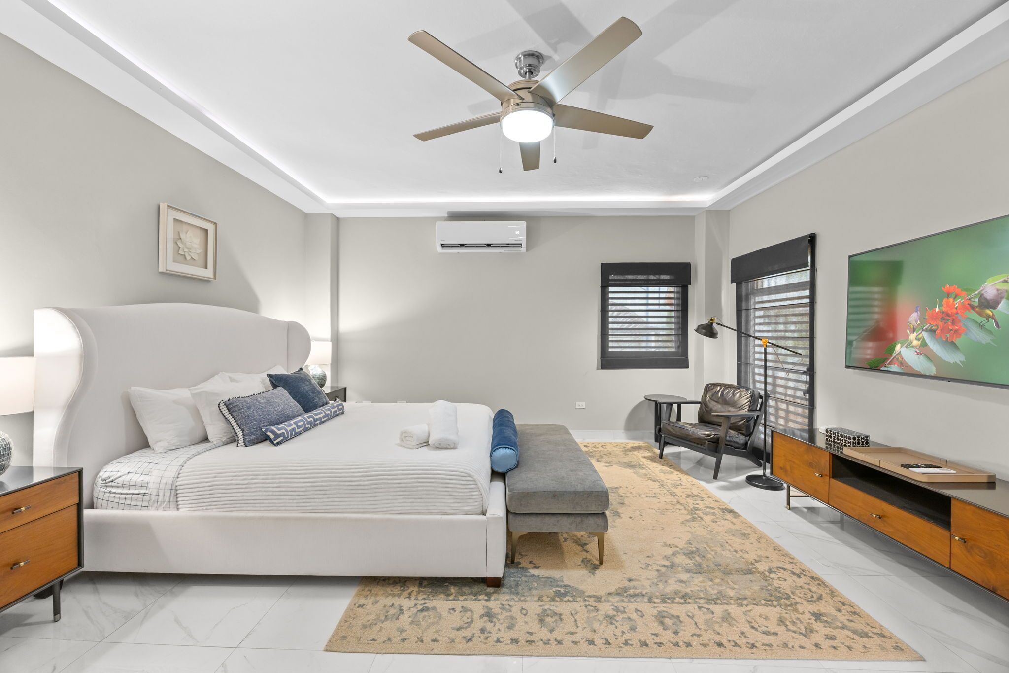 A bedroom with a bed, a ceiling fan, a TV, a window with blinds, and a small table.