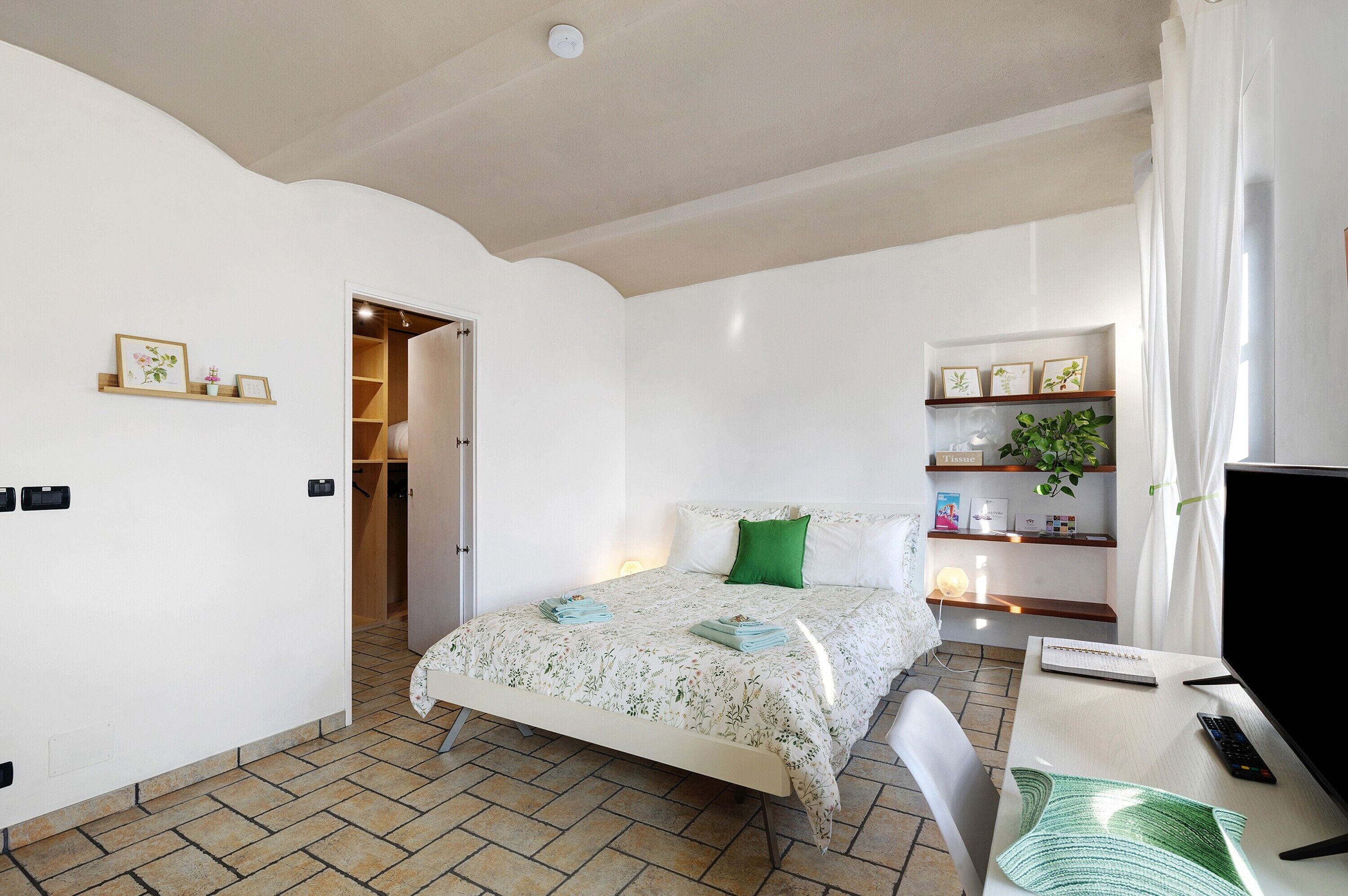 Une chambre à coucher avec un lit, un bureau, une chaise, une télévision et une étagère contenant des livres et une plante.