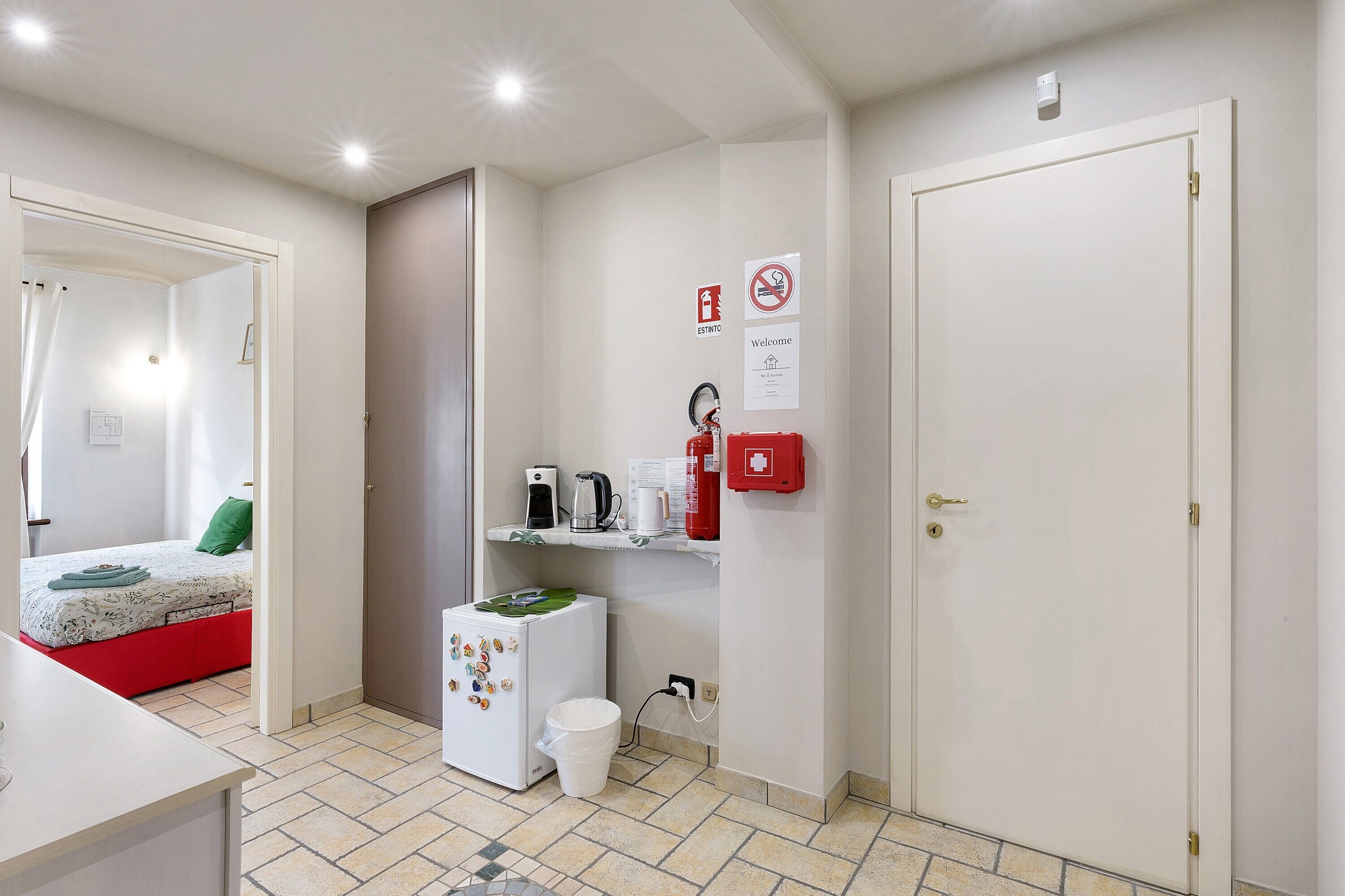 Une chambre d’hôtel avec un lit, une petite kitchenette, un réfrigérateur blanc, une poubelle blanche, une trousse de premiers secours rouge et une porte dotée d’un trou de serrure.