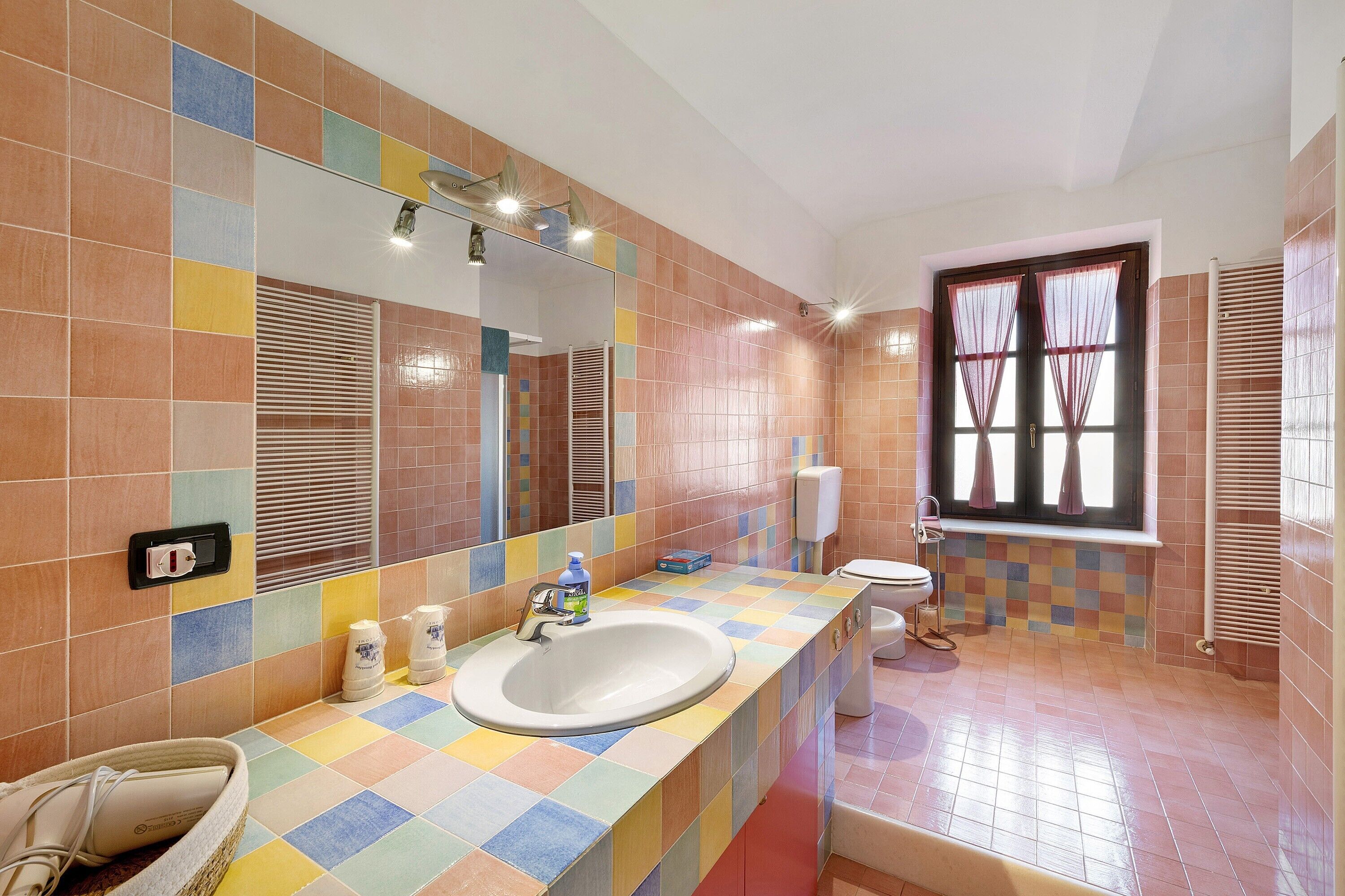 A bathroom with a tiled floor, a sink with a mirror, a toilet, and a window with curtains.