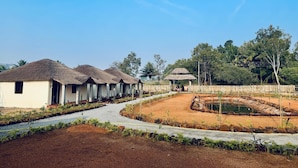 A row of thatched-roof huts with a paved pathway leading to a small pond. - Echoes Of Hampi (Koppal)