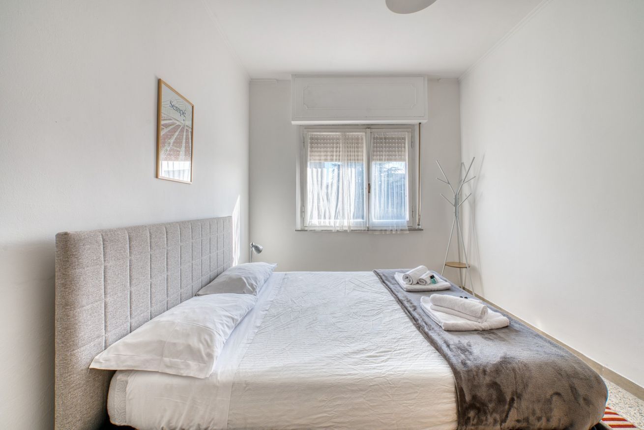 Un lit bien fait, avec une tête de lit, une table de chevet sur laquelle se trouvent un téléphone et une serviette, une fenêtre avec des rideaux et un tableau encadré accroché au mur.