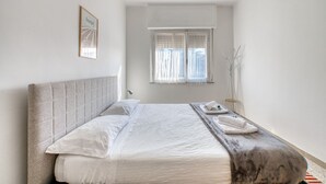 Una cama bien hecha con cabecera, una mesita de noche con un teléfono y una toalla, una ventana con cortinas y un cuadro en la pared.