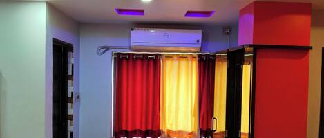 A room with a ceiling-mounted air conditioner, a curtain with red and yellow panels, and a red wall.