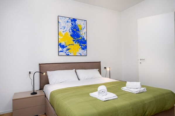Habitación de hotel con cama, mesita de noche, lámpara, colcha verde y un cuadro en la pared.