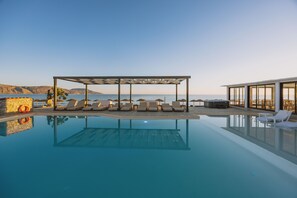 Une piscine avec des chaises longues et des parasols, un espace de détente couvert et une vue sur la mer.