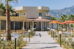 A modern building with a patio area, outdoor seating, and a mountain backdrop. - Casa di Notos Beach Hotel-Georgioupolis (Apokoronas)