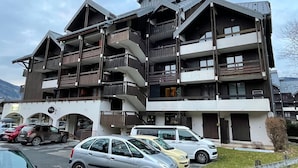 Un edificio de varios pisos con balcones, coches aparcados y montañas al fondo.