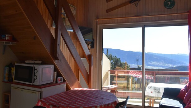 Una habitación con paredes de madera, una mesa con mantel a cuadros, microondas, un refrigerador pequeño y vistas a las montañas a través de un ventanal grande.