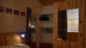 Una habitación con una cama, paredes de madera, un armario con ropa colgada y una ventana con persianas.