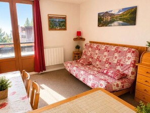 Une pièce comprenant un lit, une table avec des chaises, un radiateur, une lampe rouge, un tableau au mur et une fenêtre avec des rideaux.