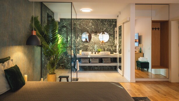 Ein modernes Schlafzimmer mit einem großen Bett, einer Zimmerpflanze und Blick in ein Badezimmer mit Marmorwand und zwei Waschbecken.