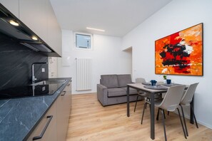 A compact kitchen with a black countertop and a grey sofa in a bright room with a large abstract painting.
