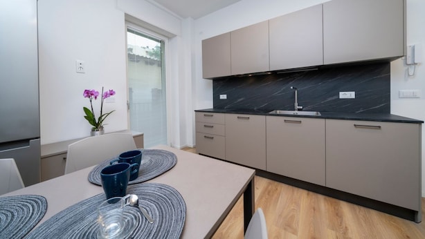 A modern kitchen with a black marble countertop, beige cabinets, and a dining area with a table set for two. - GuestHost - Cozy 60 sqm apartment, located on the first floor of a building without an elevator.The accommodation is situated in a strategic location, allowing you to reach the main points of interest within a short walking distance or by public transpor (Udine)